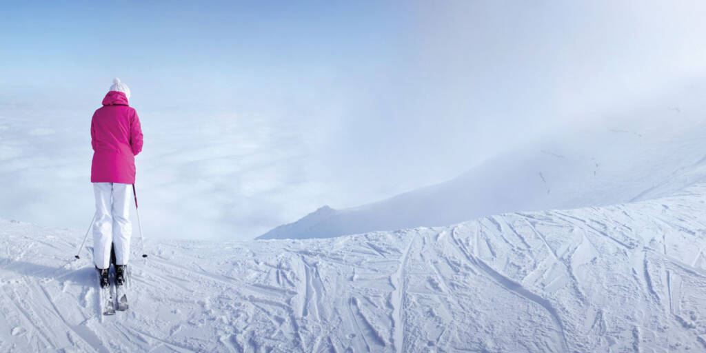 Skier in pink jacket with back to camera, looking at mountain down from top of ski run