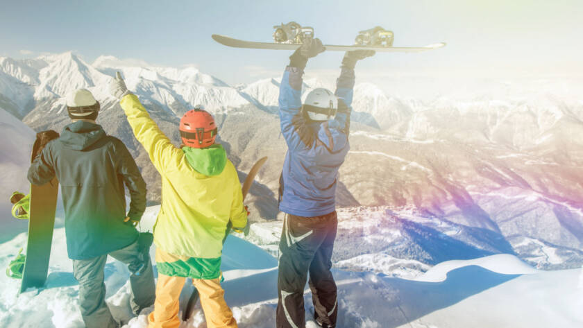 Three snowboarders looking at mountain horizon holding boards triumphantly overhead