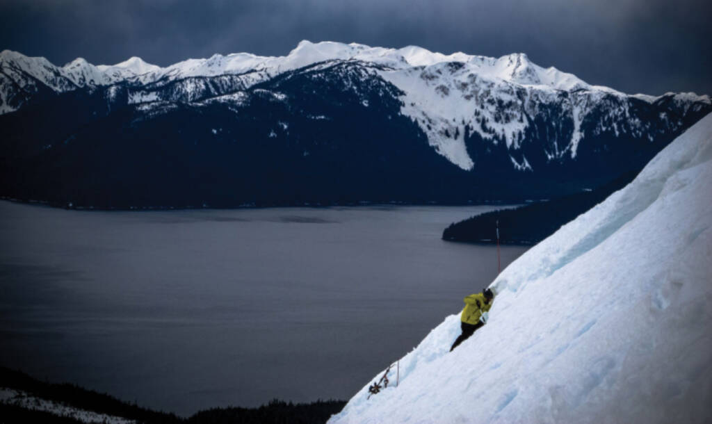 Specialist in yellow jacket installing explosives in snow in foreground, with lake and moutains in background